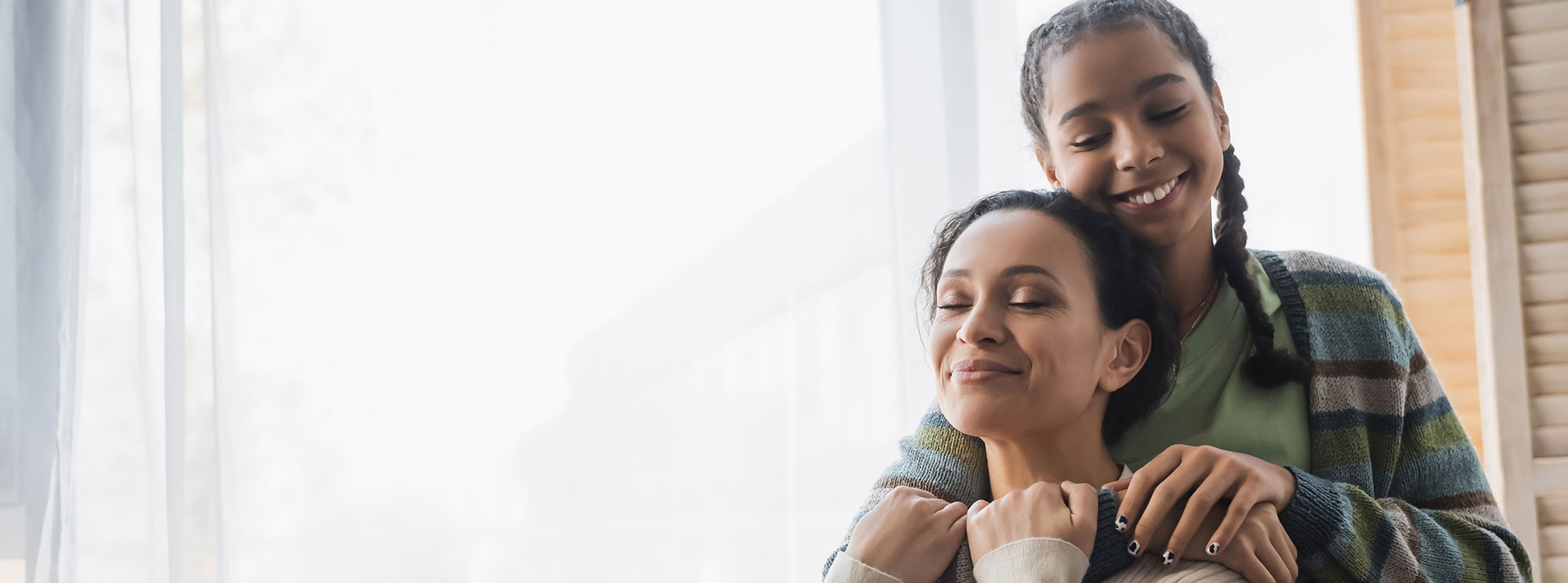 teenage african american girl hugging happy mom with closed eyes near window at home, banner